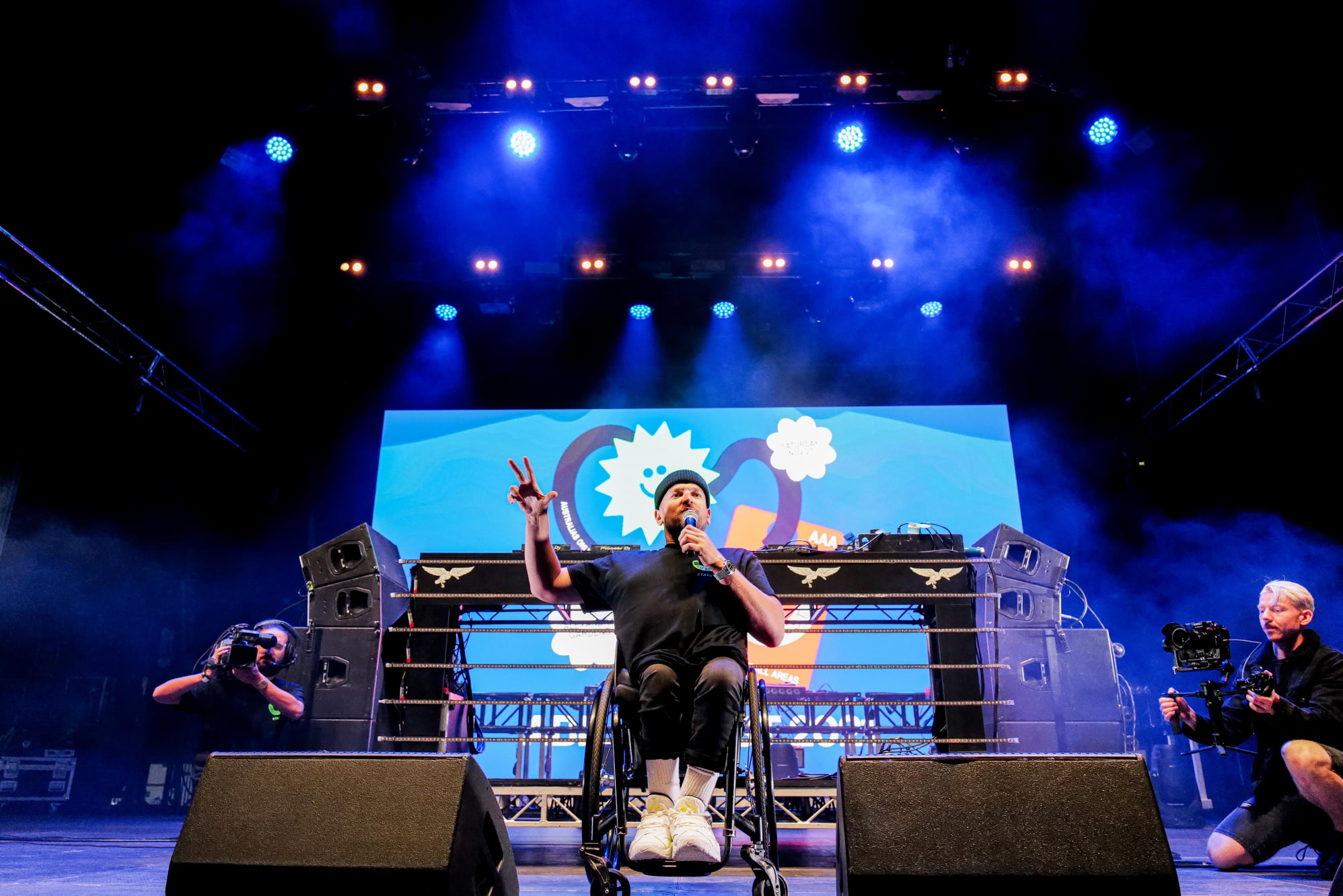 Dylan Alcott speaking on stage at Ability Fest. He is holding a microphone to his mouth and there are two photographers crouched down behind him taking photos. 
