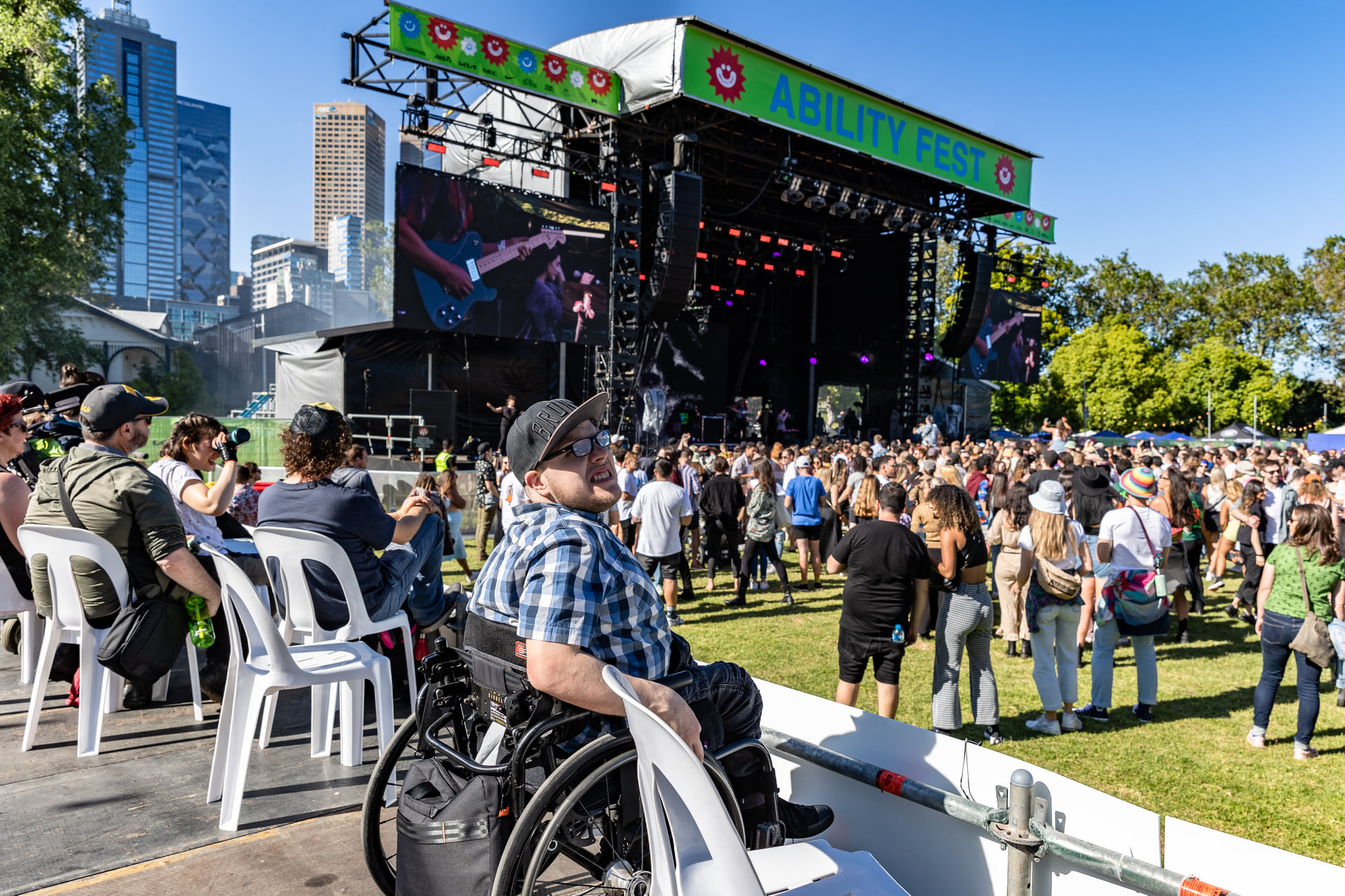 People gathering in front of an outdoor stage at Ability Fest. The sky is blue with high-rise buildings in the background. 