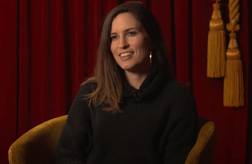 Missy Higgins, a woman with mid-length brown hair wearing all black sitting on a rust coloured velvet chair. She is smiling and looking slightly away from the camera in an interview style setting. 