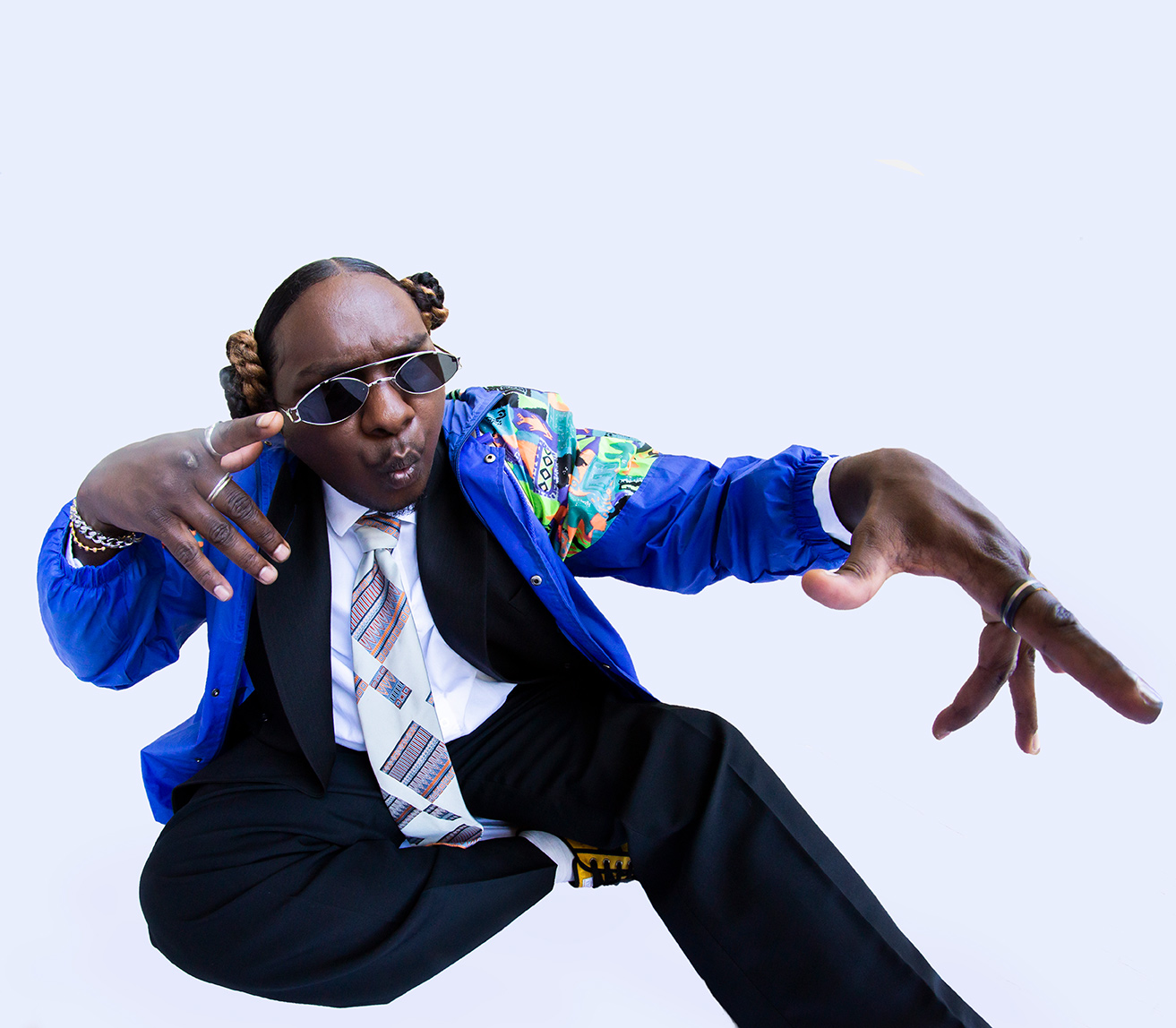 Baker Boy, dressed in a blue jacket and black trousers, his hair pulled back into two buns and sunglasses on, crouches and gestures, his mouth making an 'oo' shape, towards the camera. The photo is distorted with a fish-eye lens.