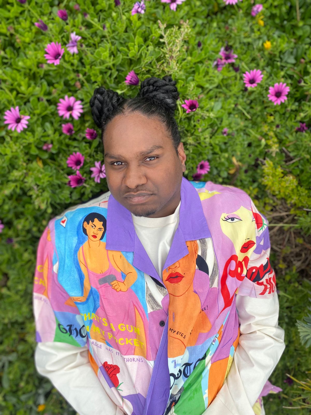 Baker Boy, his hair two buns on either side of his head, smirks at the camera. He's wearing a colourful, hand-painted jacket and lying on green grass that has flowers scattered through it.