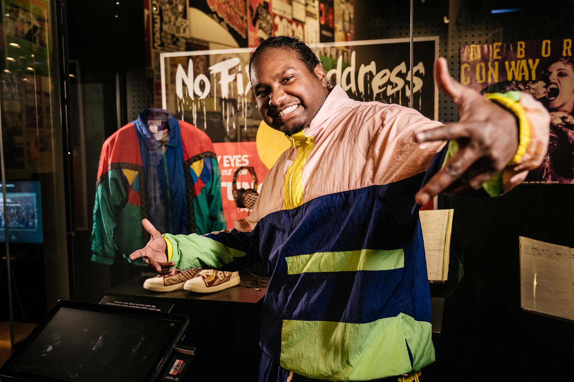 A head and shoulders of Baker Boy - a man with dark hair, tied back, wearing a tracksuit top of yellow, light pink and dark blue - standing side on to the camera, his hand out towards us and a large smile on his face. He is standing in front of a glass display cabinet in the Australian Music Vault that contains one of his jackets, a set of his headphones and a pair of his hand-painted sneakers.