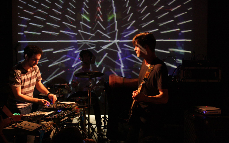 Two men stand in a dark room with a light display in the background. One man is holding a guitar, while the other uses a DJ deck. 