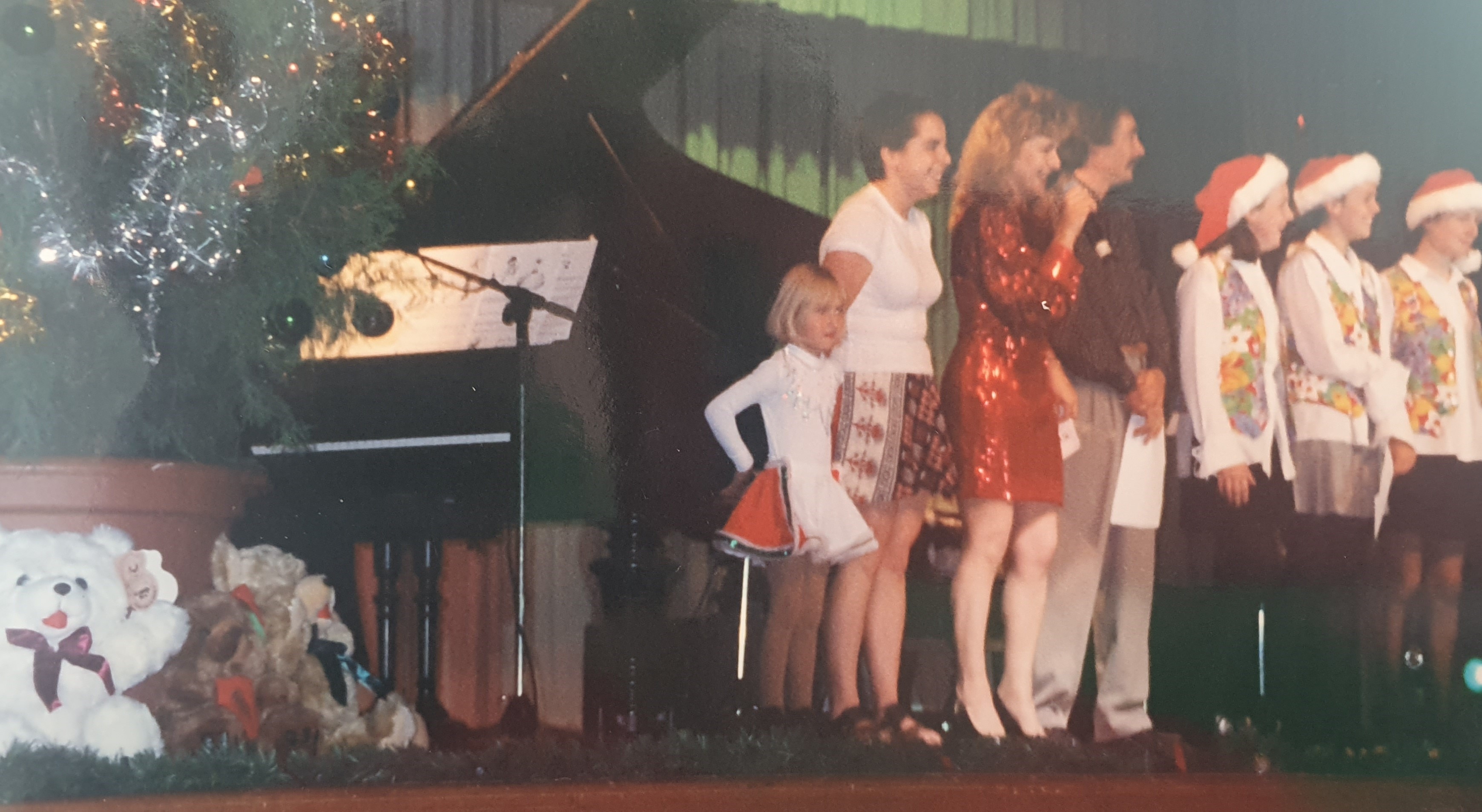 Young Alana Wilkinson and others at a Christmas concert. There is a Christmas tree and piano on stage and everyone is wearing red Santa Clause hats.