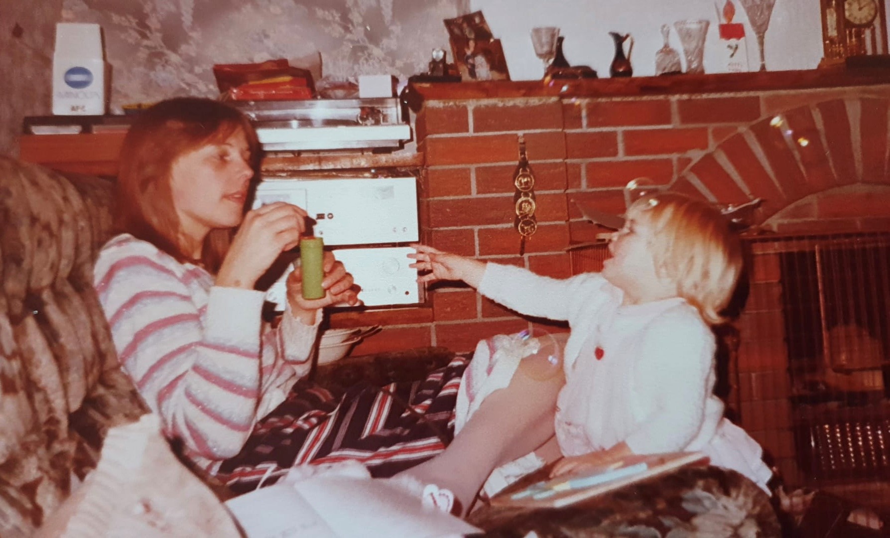 A vintage colour photograph from the 1980s showing Anna Laverty as a small, blonde child, leaning towards her mother who is sitting on a couch, covered in blankets. Her mother is holding a small, coloured tube in her hand and is talking to Anna. Behind them is a large brick mantlepiece and a silver stereo.