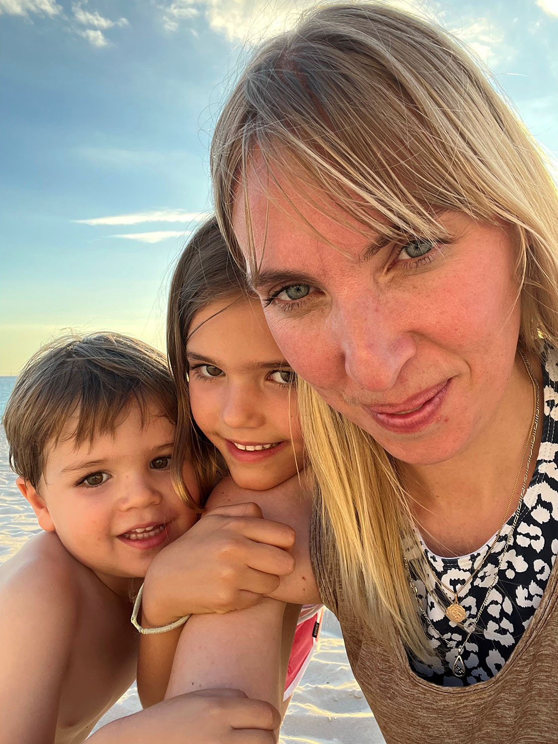 Anna Laverty, a woman with long blonde hair wearing a patterned top, takes a selfie with her two children hanging on to her arm and leaning into the camera. They're at the beach and the sand and sky are visible in the background.
