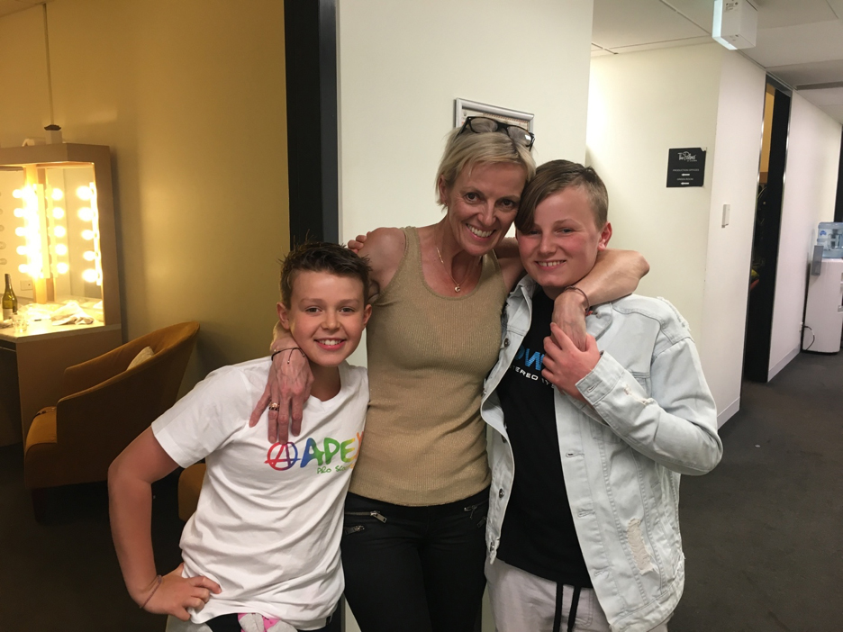 Bec Shand, a woman with short brown-blonde hair wearing a beige top and black skirt stands with her arms around the shoulders of her two sons. They are backstage and they're all smiling.