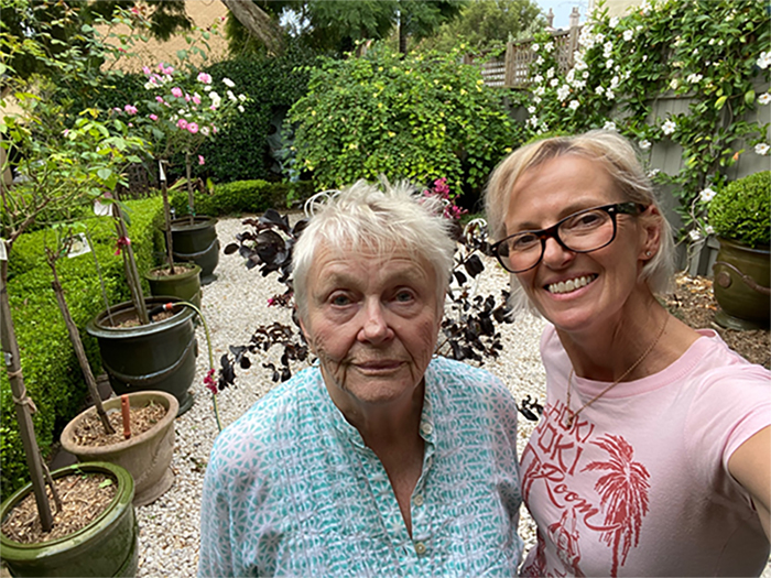 A selfie taken from above of Bec Shand, a woman with short blond hair wearing black-rimmed glasses and a pink shirt, with her elderly mother, who has short white hair and is wearing a blue gingham shirt. They are standing in what looks like a greenhouse.