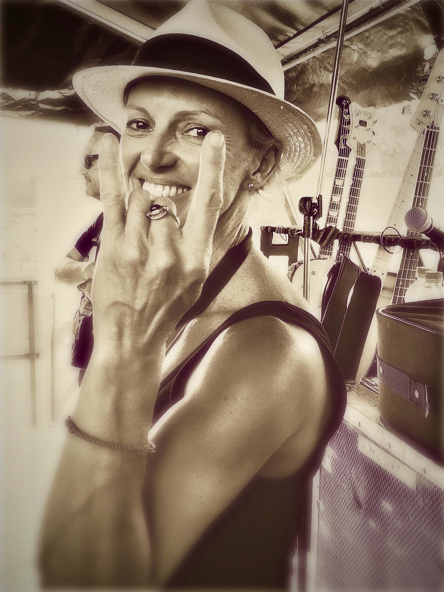 A sepia-toned photo of Bec Shand - a woman with short hair wearing a black singlet and a fedora, backstage, throwing the 'devils-horns' hand gesture that symbolises rock 'n roll.