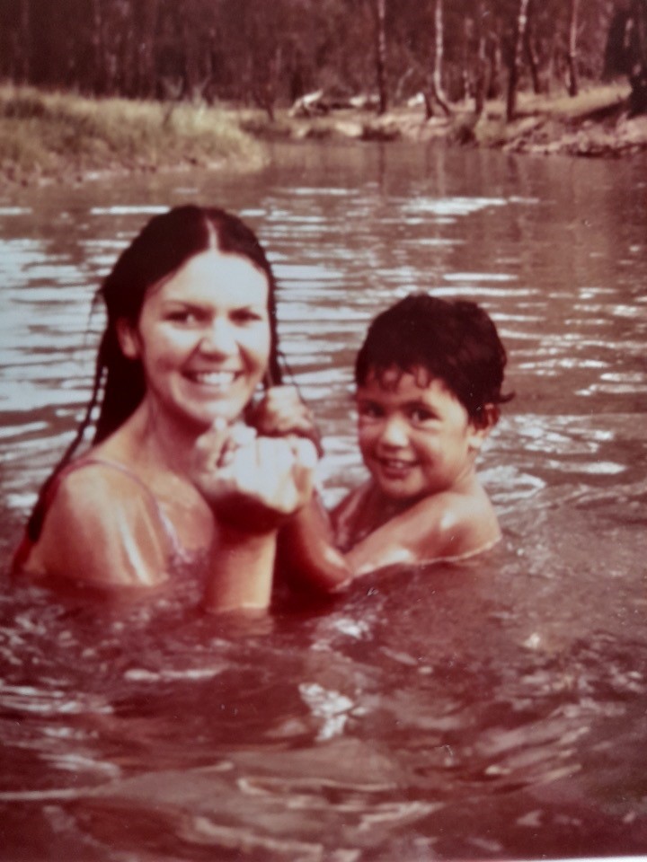 A vintage colour photograph of a woman with long dark hair, shoulder-deep in a body of water, holding the hands of a small child whose directly in front of her. They are both pictured side-on, looking around into the camera and smiling widely. The banks of the body of water are in the background, covered in grass and thin-trunked trees.