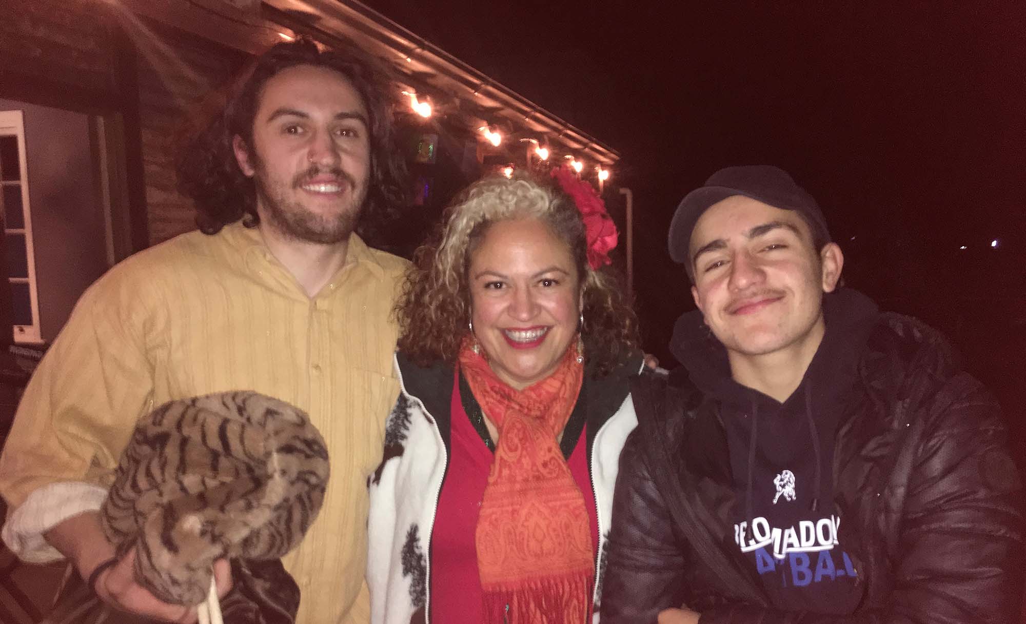 A family snapshot of Kylie Auldist - a woman with shoulder-length curly dark hair with a patch of white blonde on the top, wearing a red jumper, white and black jacket, and orange patterened scarf with a red flower tying half of her hair back - with her arms around her two sons - one standing on either side of her. They are outside at night. The son to the left is taller than her, wearing a tan shirt and carrying a cheetah-patterened jacket. The son to her right is wearing a black puffer jacket and black baseball cap. They are all smiling widely. 