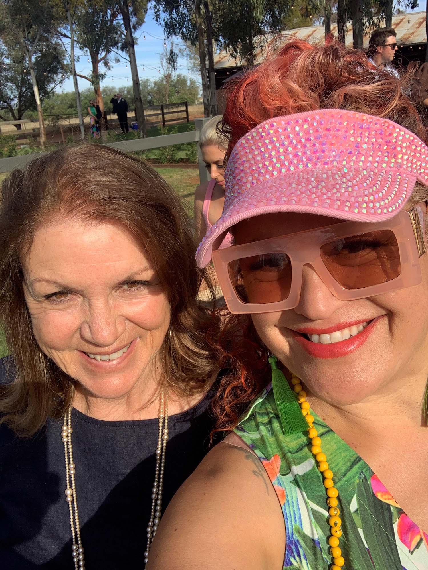 A selfie of Kylie Auldist and her mother. Kylie, to the right, is wearing a bejewelled pink visor, wide-rimmed pink sunglasses and a sleeveless green, patterened top. Her mother, posed slightly behind Kylie, has shoulder-length brown hair and is wearing a dark blue top and a double stranded necklace. They are both standing outside during the day, and smiling widely.