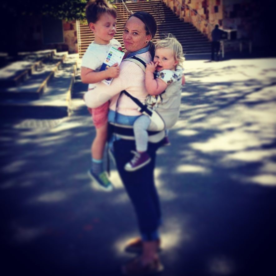 A mother standing in a street with a child in a carrier on her back and one in her arms. 