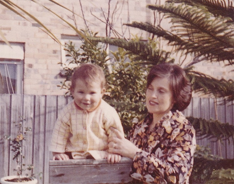 A vintage, textured colour photo of a woman in a backyard, holding a toddler, who is perched on a fence post.