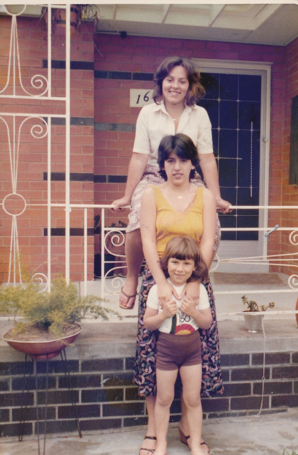 A vintage colour photo of three girls in front of a suburban house - each stands in front of the other, the tallest at the back and the smallest at the front, forming step stairs of sisters.
