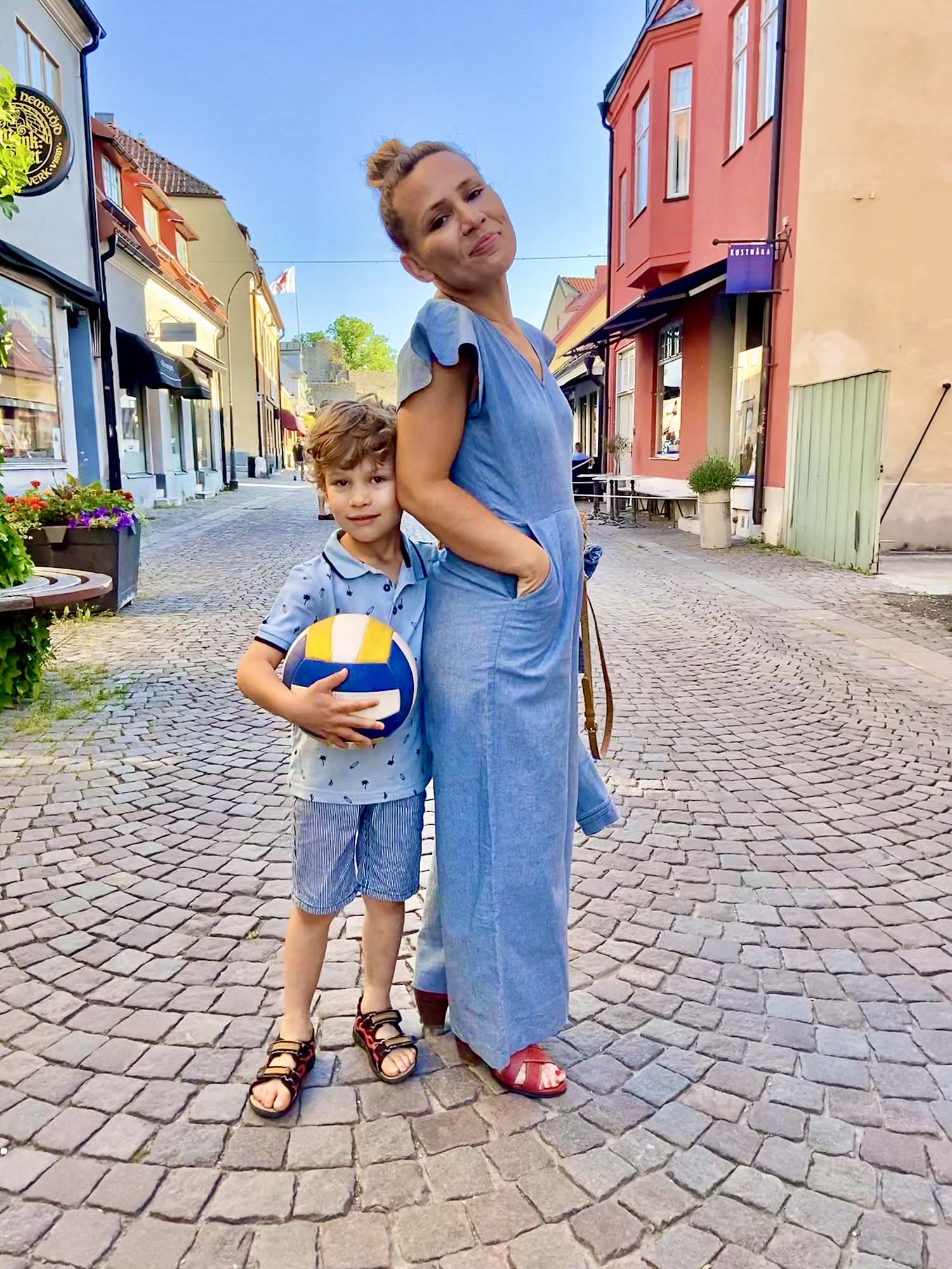 Tania Doko, a woman with blonde hair that's tied up, wearing a blue jumpsuit, stands side on to the camera, looking over her shoulder at it and smiling cheekily. Clinging to her back is her son - a small boy wearing a blue polka-dot shirt and denim shorts and carrying a ball.