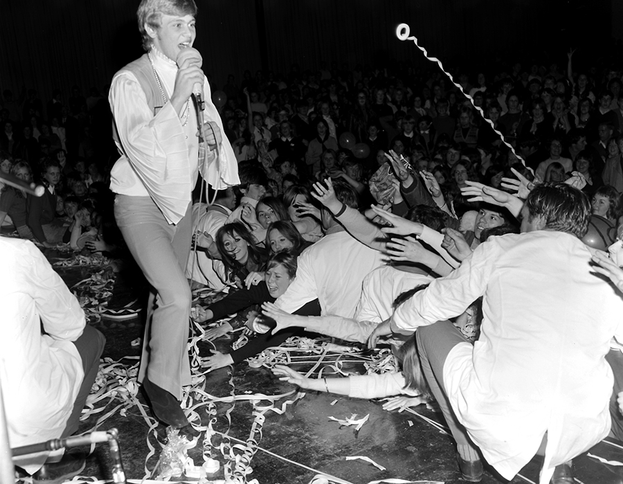 A black and white image of John Farnham performing on stage at Festival Hall in August of 1969. John is wearing flared pants, boots and a long sleeve shirt with flared sleeves and a vest. He is holding a microphone to his mouth, as the crowd reaches for him and throws streamers onto the stage.