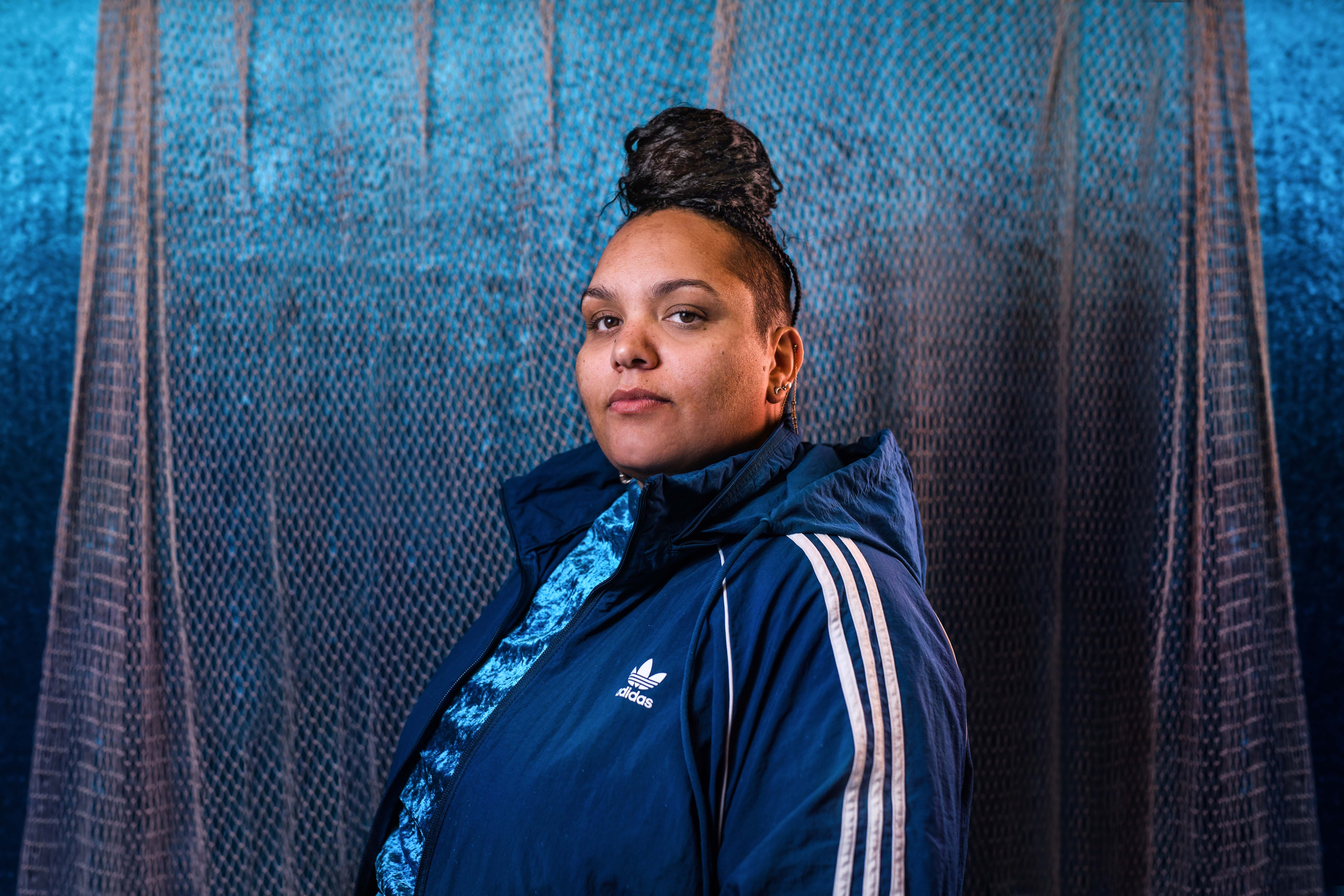 Lady Lash stands side on, looking directly into the camera. She wears a blue, crush velvet top under a navy Adidas jacket. Her hair is gathered into a large bun on the top of her head and she wears minimal makeup. Behind her a cream, mesh curtain covers a blue velvet wall.