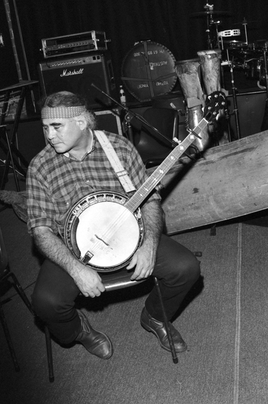 A black and white photo taken from above of Kev Carmody sitting on a stool, a banjo slung across his body. He's wearing a checkered shirt and jeans and looking off toward the side. He appears to be inside a bar.