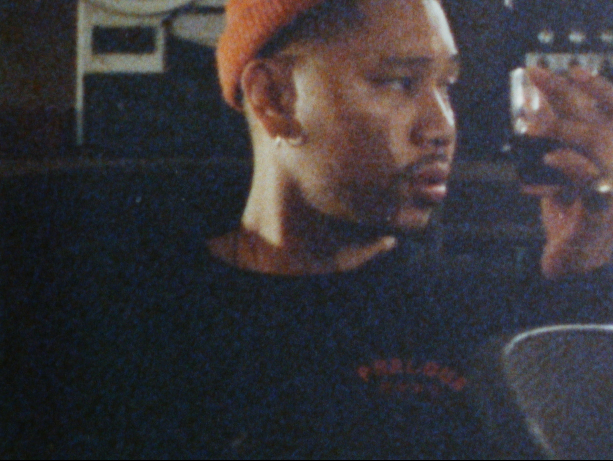 A faded and grainy close-up of Silent Jay, a man wearing an orange beanie and a dark shirt. He's sitting down inside and looking towards the side, holding up a drink in his hand.