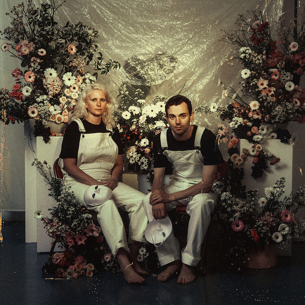 The two members of Big Scary, dressed in white overalls and black t-shirts, sit within an elaborate set of fake flowers, plinths and a fabric backdrop. The image has been treated to look grainy and dim, like a vintage photo.