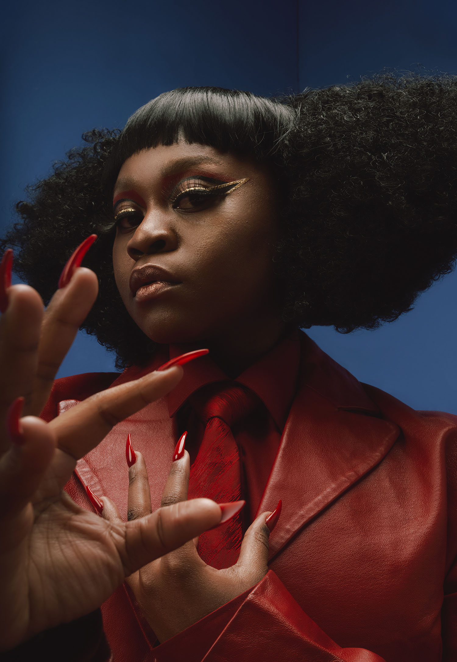 A dramatically lit portrait of Sampa the Great. Their hair is bunched to both sides, they're wearing an orange jacket. One hand is to their heart, the other stretched close to the camera, the fingers tipped in long red fingernails. The background is blue.
