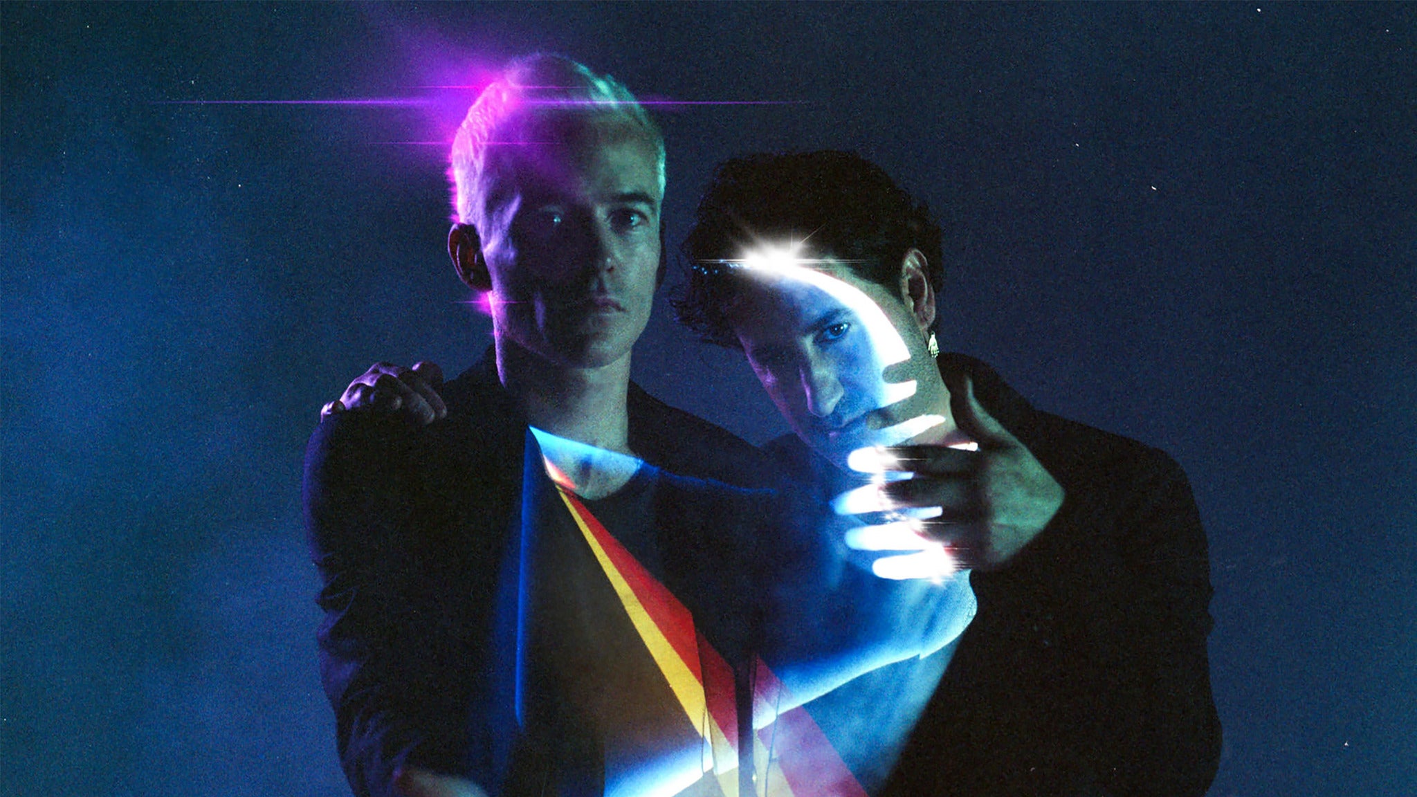The two members of The Avalanches, shown from the waist up against a dark, mottled blue background. There's a bright pink highlight behind one of their heads, and there is what looks like a prism in front of them, one side showing yellow and red, and the other emitting a line of white light that picks out the other's face.