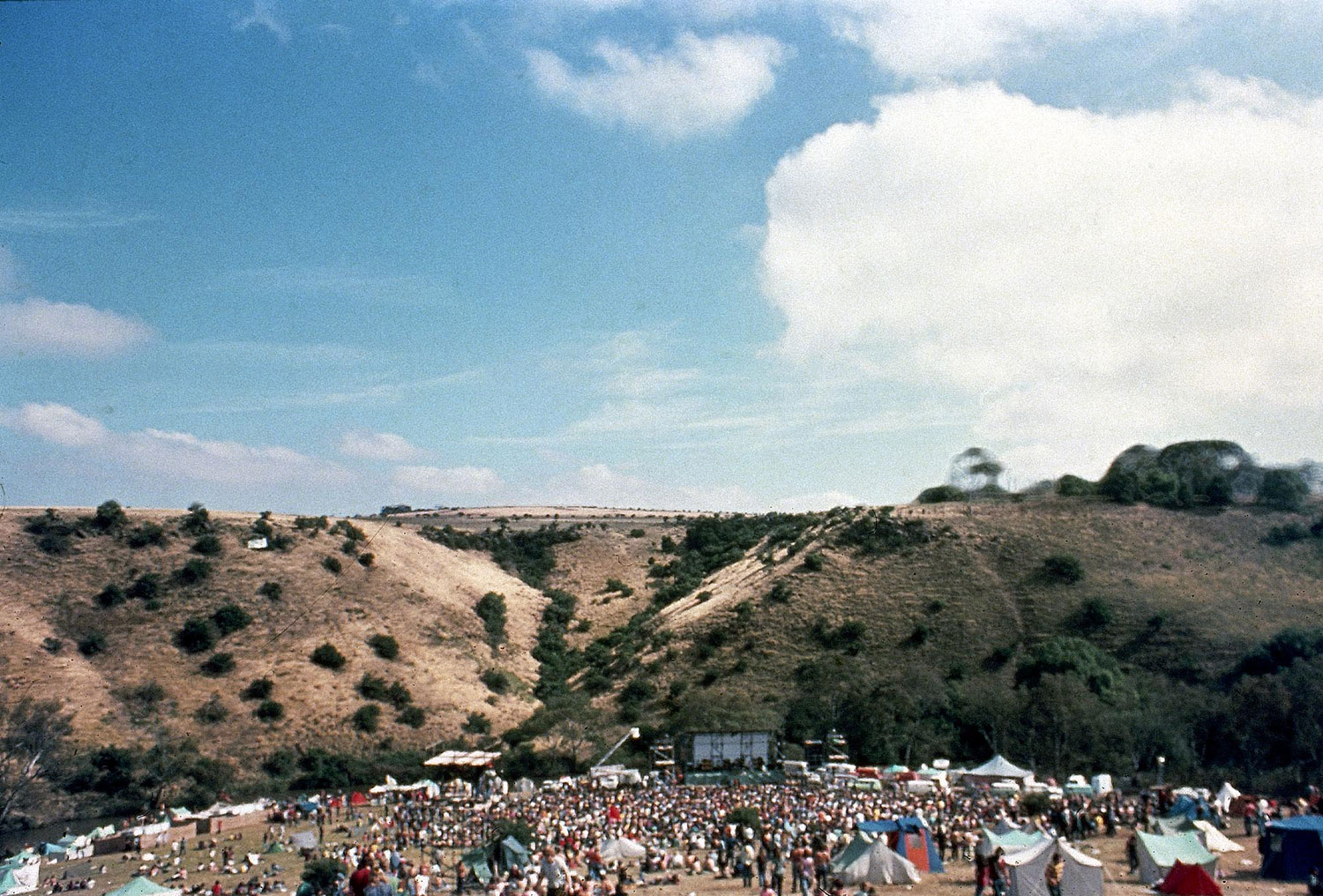 A vintage colour photo of the Sunbury Music Festival site, taken from the sky, showing a large crowd in a valley, with hills in the background and blue sky.