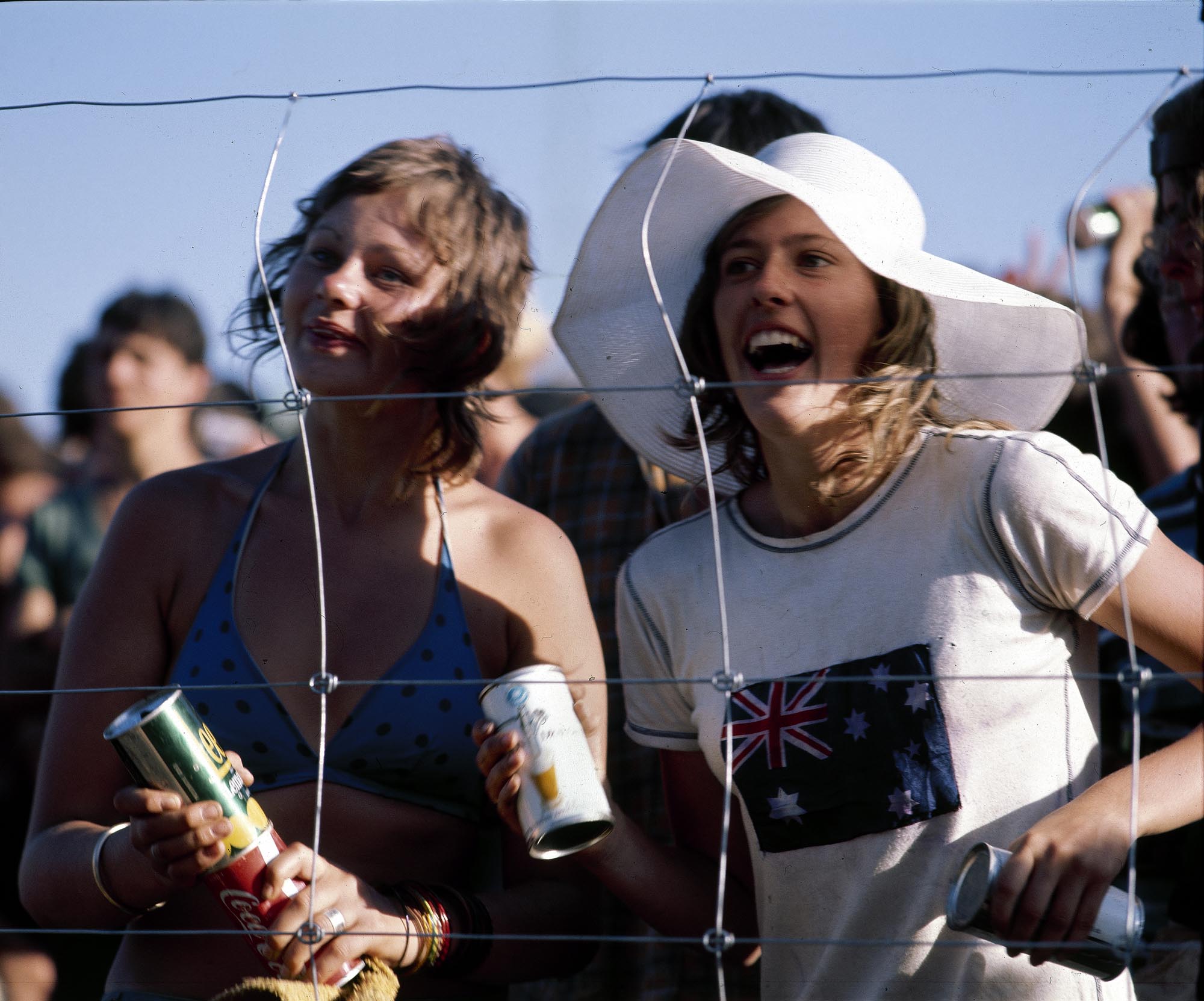 A vintage colour photo of two young women watching the Sunbury Music Festival. They are shown from the waist up, holding cans of beer and enjoying themselves. One is wearing a large white sunhat. Behind them are more audience members.