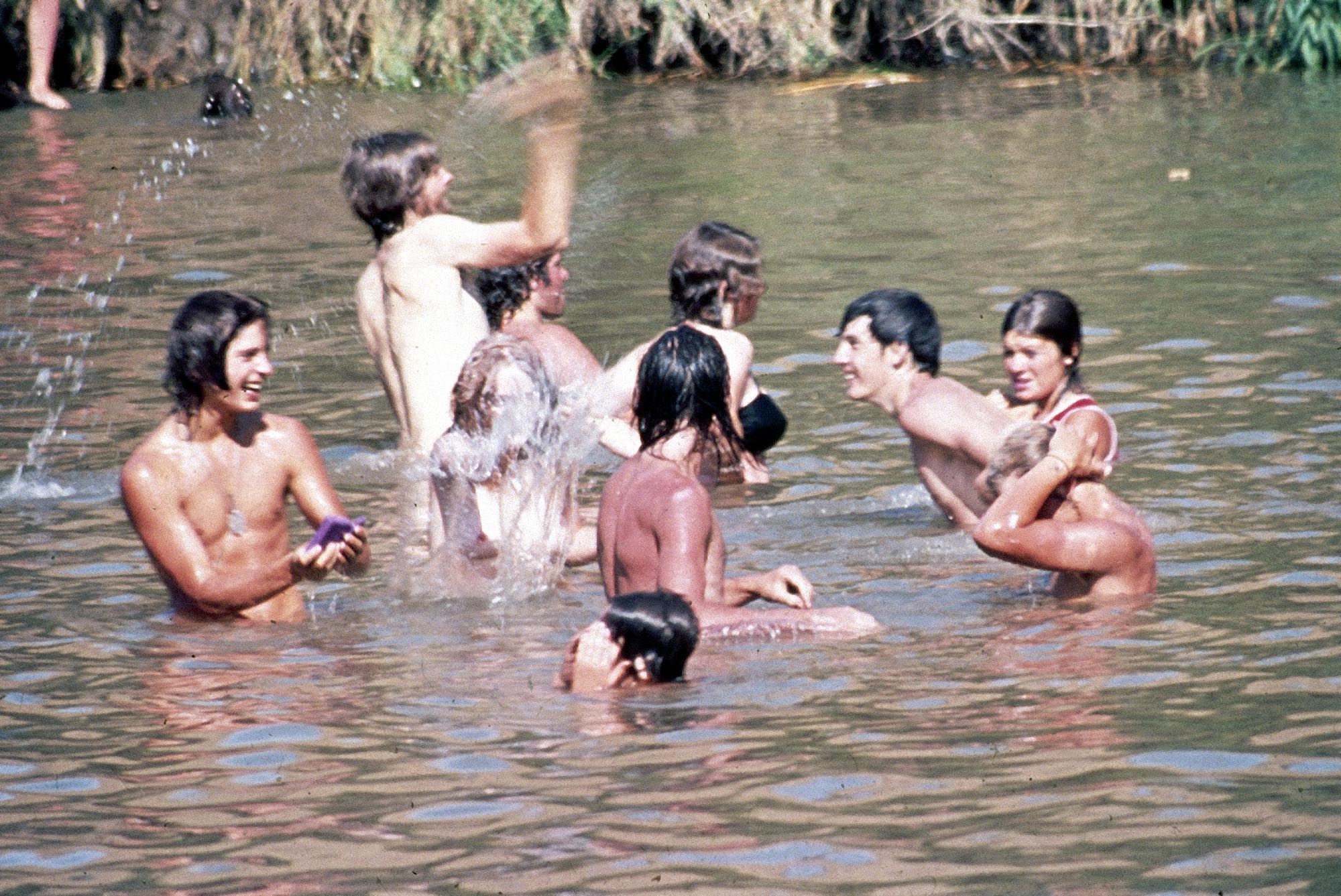 A vintage colour photo of a group of young people playing in what appears to be a dam, with water up to the waists of some and the shoulders of others.