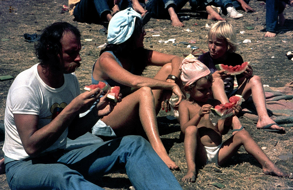 A vintage colour photo, taken from the side, showing a man, a woman and two small children sitting on the grass eating watermelon at the Sunbury Music Festival. It appears to be a very hot day and the woman has a shirt wrapped around her head for sun protection.