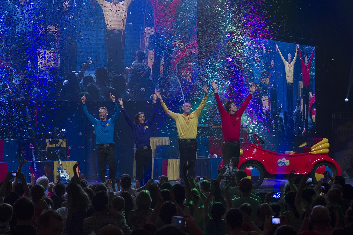 The four original Wiggles perform live onstage in a darkened space. They are all holding hands, their arms straight up in the air as if about to take a bow. To their right is the Big Red Car and behind them are foldable video screens show an enlarged picture of them. In between the video screens and The Wiggles, large glitter cannons go off, throwing multicoloured glitter into the air. In the foreground a large audience of children and adults enjoy the show.