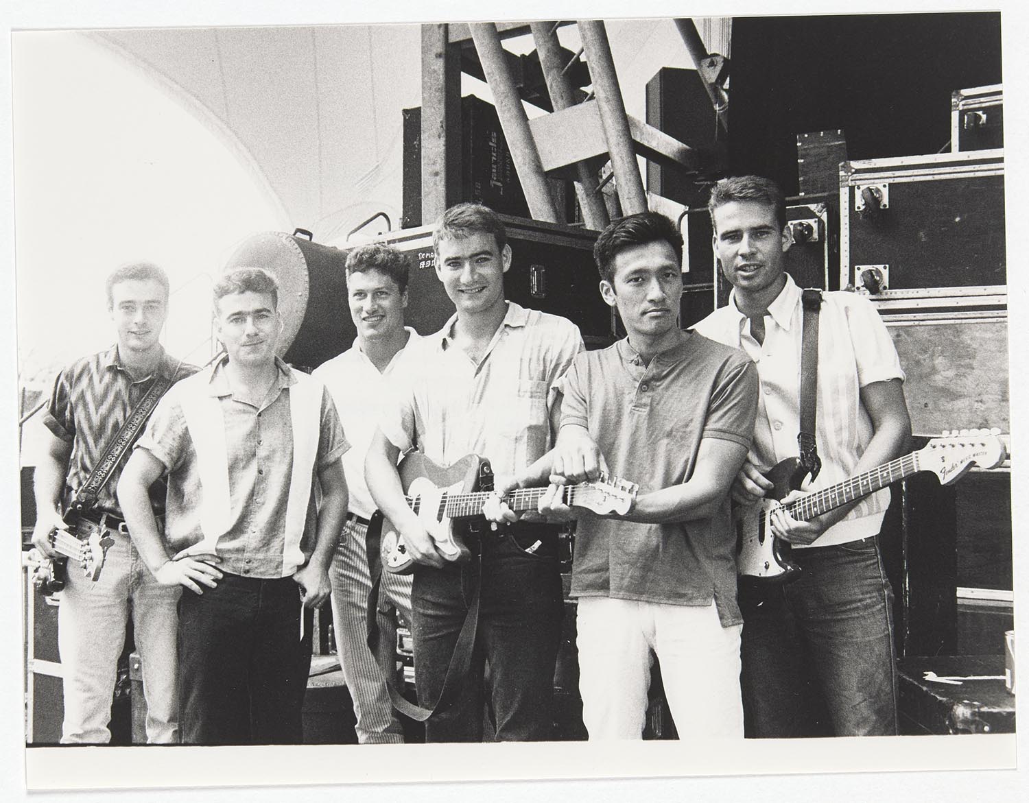 A black and white photo of the band The Cockroaches, posing in front of road cases at the side of a large outdoor stage. They're all wearing casual clothes and two of them have guitars slung around their necks.