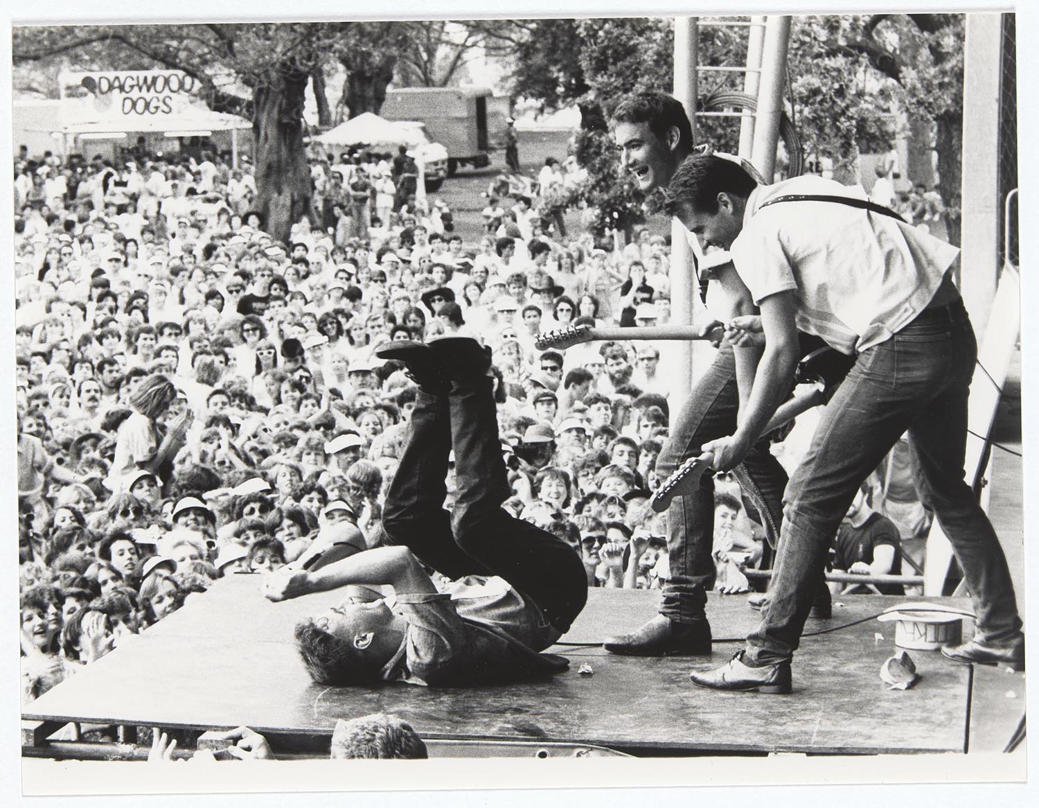 A black and white photo of the band The Cockroaches performing on a large outdoor stage, shown from side of stage. One band member is on their back, their hands and feet up in the air like a dead bug, while two other band members lean over them, playing guitar and smiling. In the background a large crowd enjoys the show.