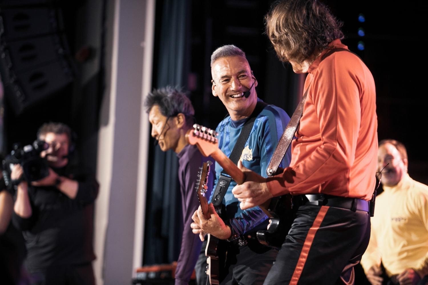 The four original Wiggles perform live on stage, smiling happily. In the background, a cameraman films them.